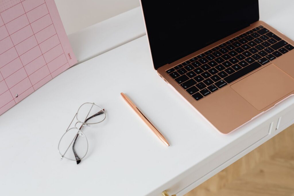 An open laptop on a clean white desk beside a notebook and glasses.